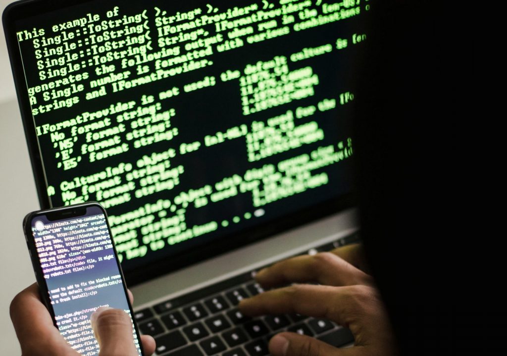 A hooded figure engaged in hacking using a laptop and smartphone in low light.